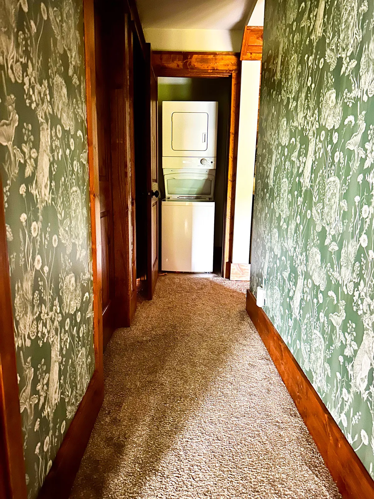 A functional yet inviting laundry area features modern laundry appliances against decorative wallpaper and plush carpeting. The space is bright and organized, supporting the comfort and convenience promised to guests.