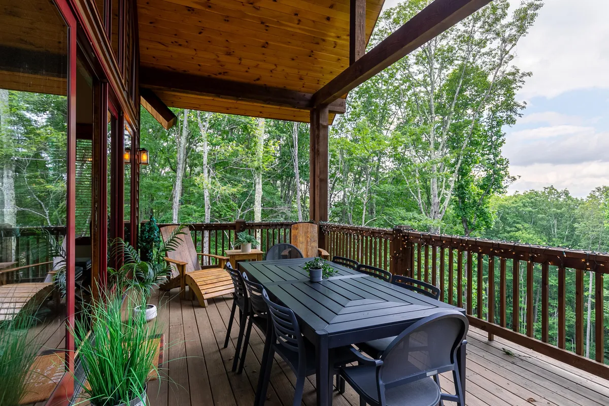 An inviting outdoor dining table surrounded by wooden chairs, set on a deck with glass railing and lush greenery all around. The scene suggests tranquil outdoor meals with expansive views into the mountains, ideal for sunset dinners or stargazing. Soft, ambient lighting enhances the evening mood.