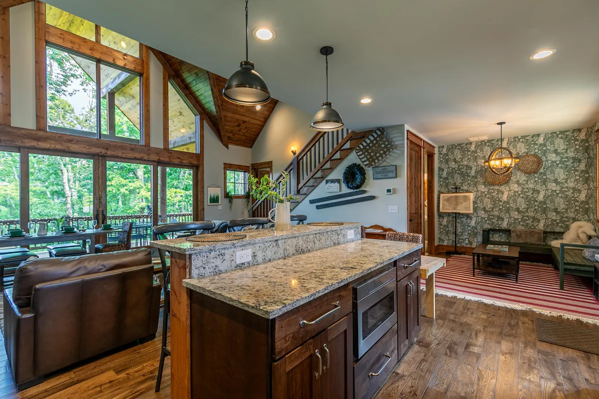 An open family room with expansive windows offering scenic views, a cozy seating area, an island countertop perfect for casual breakfasts, and a staircase adorned with tasteful decor. The space exudes warmth and sophistication, with contemporary furnishings and rustic touches.