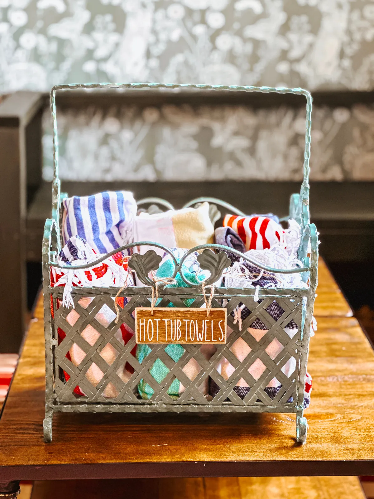 A playful interior detail featuring a metal basket filled with towels next to a sign reading 'HOT TUB TOWELS.' The vignette evokes relaxation and enjoyment, suggesting the presence of a private hot tub amenity for guests to unwind in after a day of exploring.