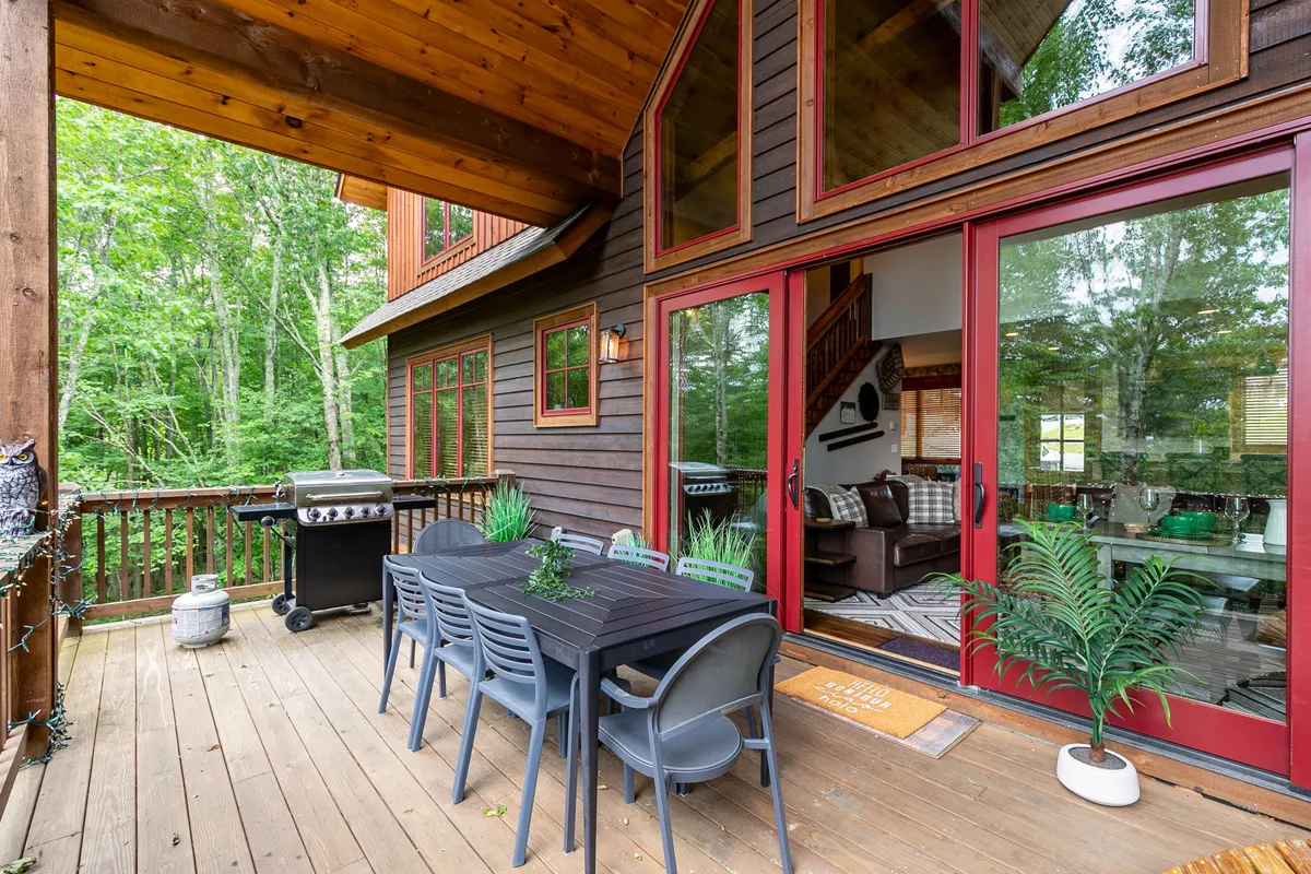 This image features an inviting outdoor dining table positioned on a spacious deck, complemented by a propane grill and surrounded by lush plants. The scene is set for an alfresco meal, with large windows reflecting the greenery and a cozy atmosphere that suggests both relaxation and social gatherings.