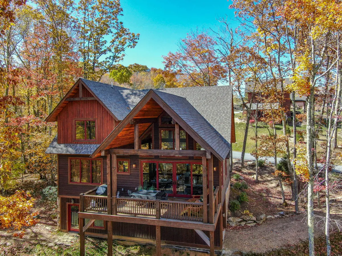 A broad wooden deck attached to the home, bordered by large windows and framed by colorful autumn foliage and sturdy trees. The space is set up for gatherings or relaxation, with nature closely surrounding the deck and home. The view highlights the seamless blend between indoor comfort and outdoor beauty.