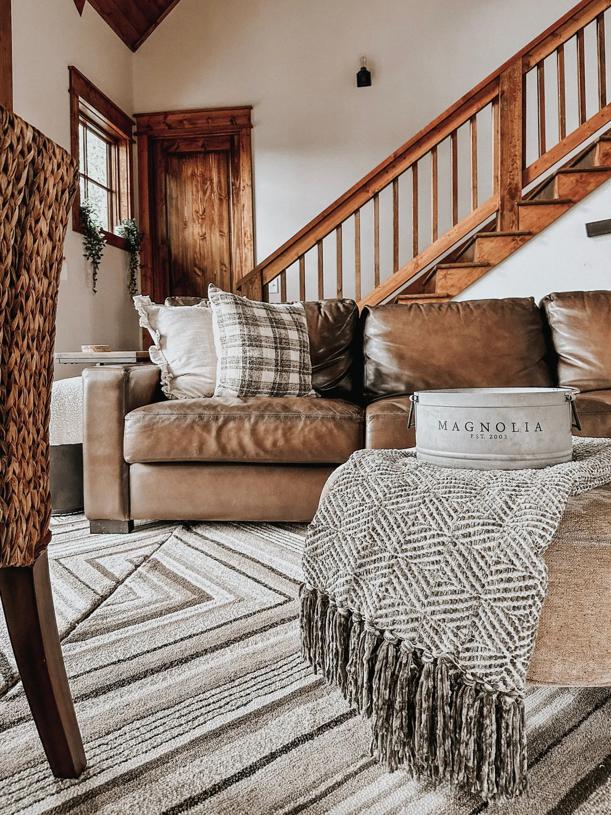 A cozy loft corner with a leather sofa adorned with plaid pillows and a woven chair arranged for conversation or quiet reading. The staircase hints at the upstairs setting, while a decorative blanket and bucket add rustic charm. The warm, welcoming setup is perfect for adults seeking a peaceful retreat within the playful loft environment.