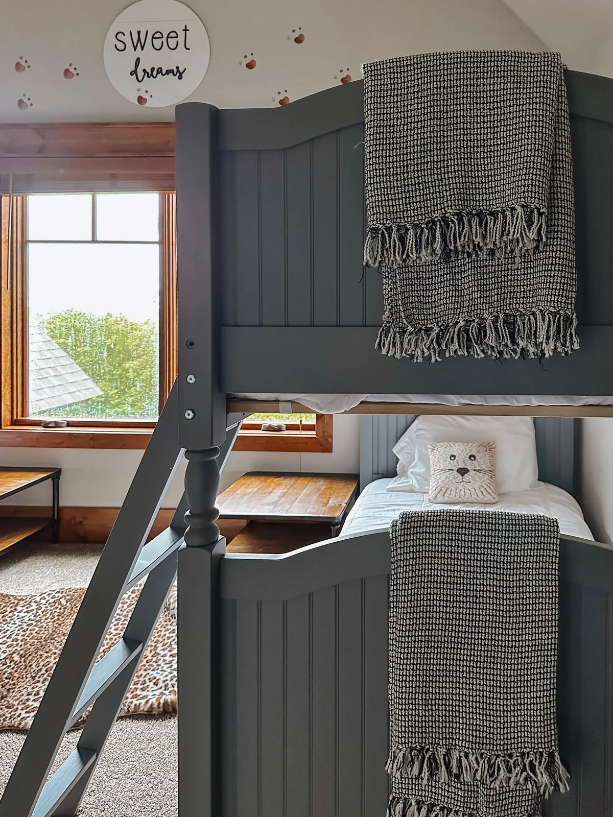 A playful bunk room features sturdy wooden bunk beds adorned with plaid blankets and whimsical owl pillows. Large windows let in ample light and offer views of the surrounding outdoors, while a 'sweet dreams' sign adds a cozy, welcoming touch.