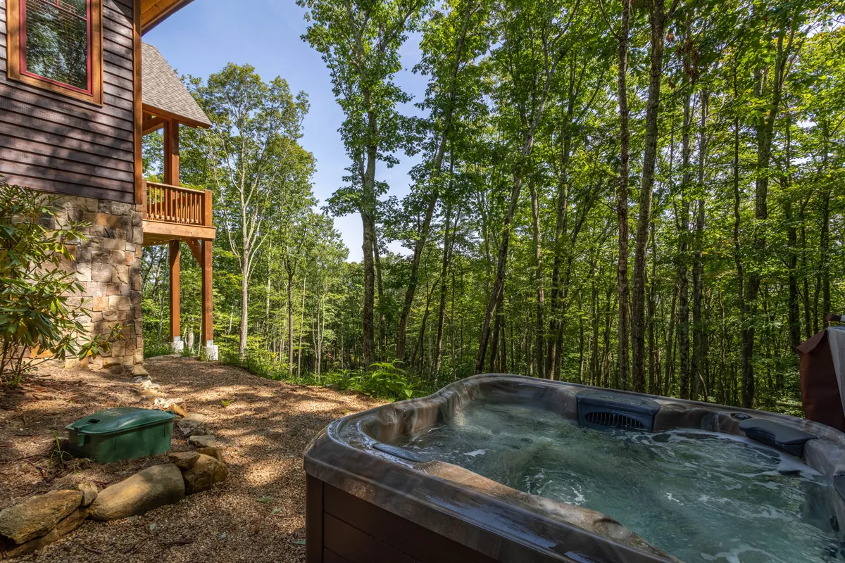 A serene outdoor hot tub is nestled on the deck, surrounded by mature trees, offering an intimate spot for soaking while appreciating the beauty of nature. The house and deck are visible, reinforcing the connection between indoor comfort and outdoor luxury.