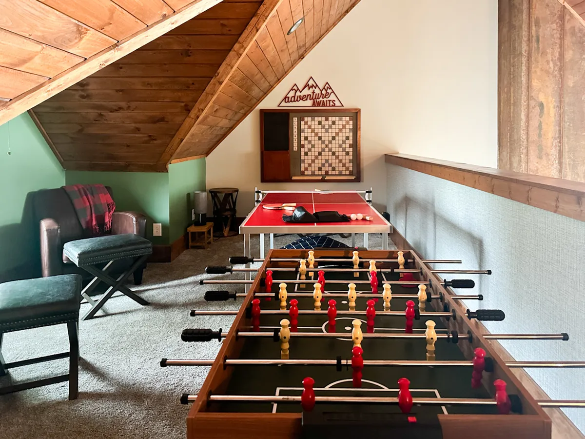 A lively loft space centered around a foosball table and a versatile game table, complemented by a welcoming seating area. The room features a wooden ceiling and mountain-themed decor, exuding a playful and relaxed vibe. The arrangement invites both competitive play and group gatherings, with ample space for board games and casual lounging.