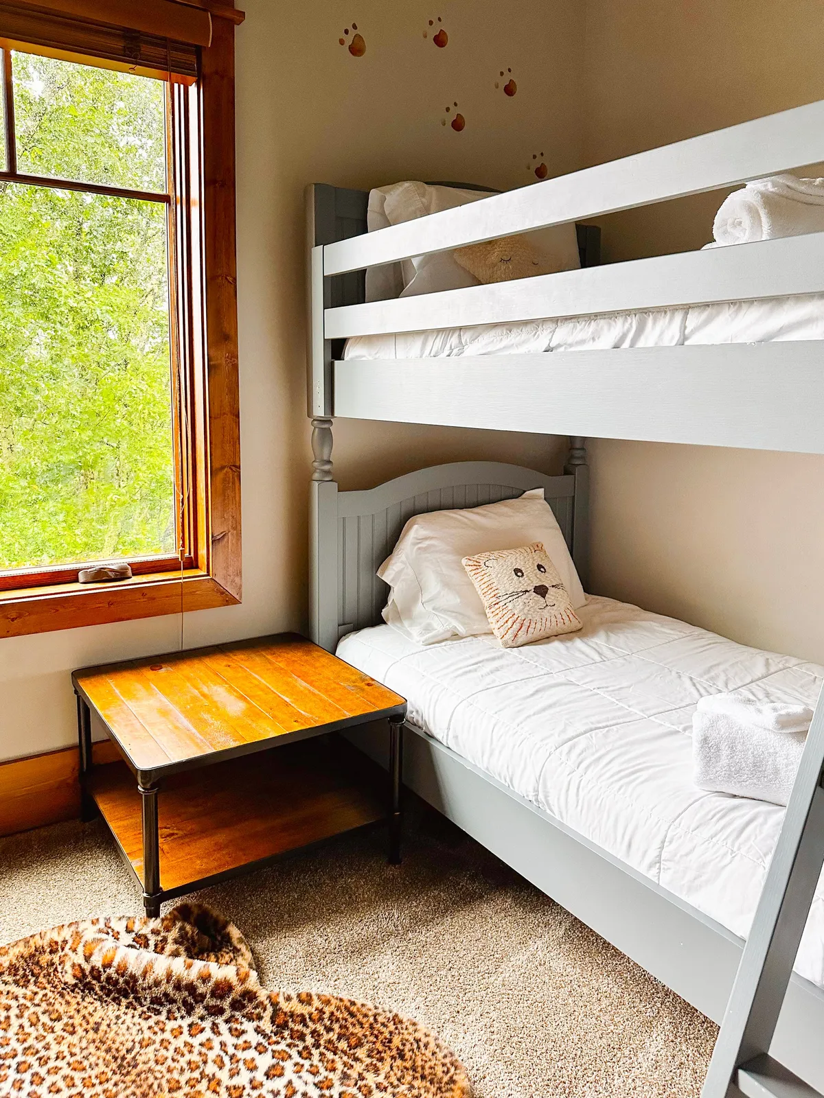 A playful, inviting kids' bunk room with wooden bunk beds, a window overlooking greenery, a wooden side table, and an animal-themed pillow. The space radiates warmth and the potential for fun, storytelling, and imaginative play.