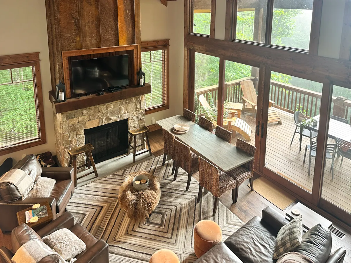 A spacious open-concept living and dining area featuring a striking stone fireplace, floor-to-ceiling windows framing panoramic mountain views, a large dining table for family meals or gatherings, and an inviting seating area with comfortable sofas and armchairs. Natural light floods the space, highlighting the blend of rustic charm and modern comfort.