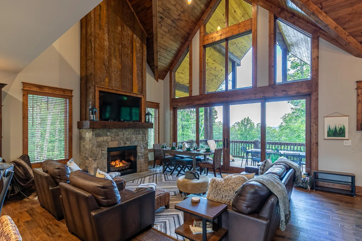 The image showcases a stunning, open-concept living and dining area with expansive windows framing mountain views, a grand rustic stone fireplace, plush leather sofas for cozy gatherings, and an elegantly set dining table—blending comfort and sophistication in a mountain retreat setting.
